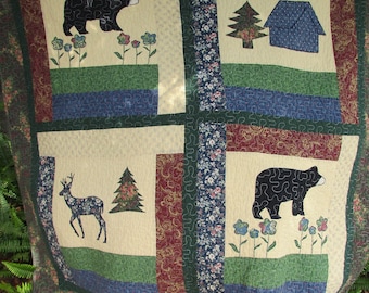 May include: A beige and green quilt with a patchwork design featuring black bears, deer, trees, and houses. The quilt has a blue and red border.