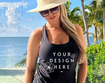 May include: Black tank top with the text "YOUR DESIGN HERE" and a floral graphic. The person is wearing a straw hat and sunglasses. The background includes a view of the ocean and palm trees.