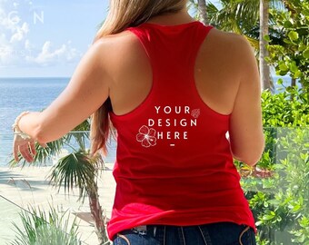 May include: A red racerback tank top with the text "YOUR DESIGN HERE" and a floral graphic. The tank top is paired with denim shorts. The scene is set on a balcony overlooking the ocean, with palm trees in the background.