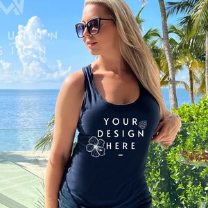May include: Navy blue tank top with the text "YOUR DESIGN HERE" in white. The tank top is paired with denim shorts. The background features a tropical beach scene with palm trees and a clear blue sky.