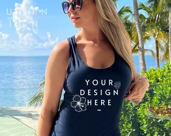 May include: Navy blue tank top with the text "YOUR DESIGN HERE" in white. The tank top is paired with denim shorts. The background features a tropical beach scene with palm trees and a clear blue sky.