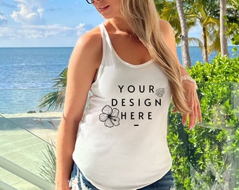 May include: White tank top with the text "YOUR DESIGN HERE" and a floral graphic. The model is wearing a straw hat and sunglasses. The background features a tropical beach scene with palm trees and ocean views.