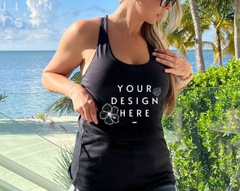 Puede incluir: Camiseta sin mangas negra con el texto "YOUR DESIGN HERE" y un diseño floral. La camiseta sin mangas se usa con pantalones cortos de mezclilla. El fondo presenta una escena de playa tropical con palmeras y cielo azul.