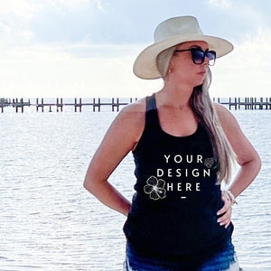 May include: A woman wearing a black tank top with the text "YOUR DESIGN HERE" and a floral graphic, paired with denim shorts. She is wearing a wide-brimmed hat and sunglasses, standing on a sandy beach with the ocean in the background.