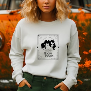 May include: White sweatshirt featuring a black graphic with the text "It's just a bunch of Hocus Pocus." The graphic includes silhouettes of three witches and decorative border elements. The sweatshirt is a long-sleeved crew neck.