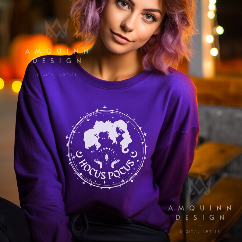 May include: A purple sweatshirt featuring a white graphic of three witch silhouettes within a circular design, with the words "Hocus Pocus" in a circular pattern. The sweatshirt is a solid purple color.