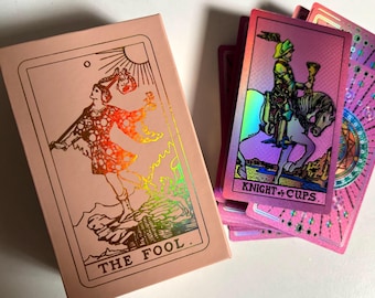 May include: A pink tarot card deck with holographic foil accents. The box features an illustration of a figure labeled "THE FOOL." Several cards are visible, including one labeled "KNIGHT of CUPS," with a knight on horseback. The deck has a mystical theme.