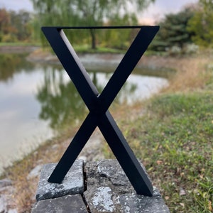 May include: Black metal table legs in an X shape, with a triangular top. The legs are standing on a stone surface, with a pond and trees in the background.