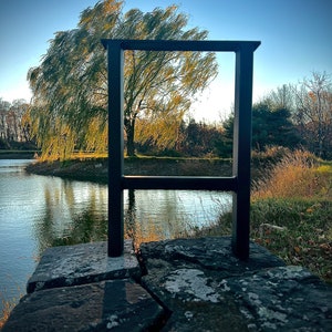 May include: A black metal frame stands on a rocky surface near a pond. A weeping willow tree with green leaves is visible through the frame.