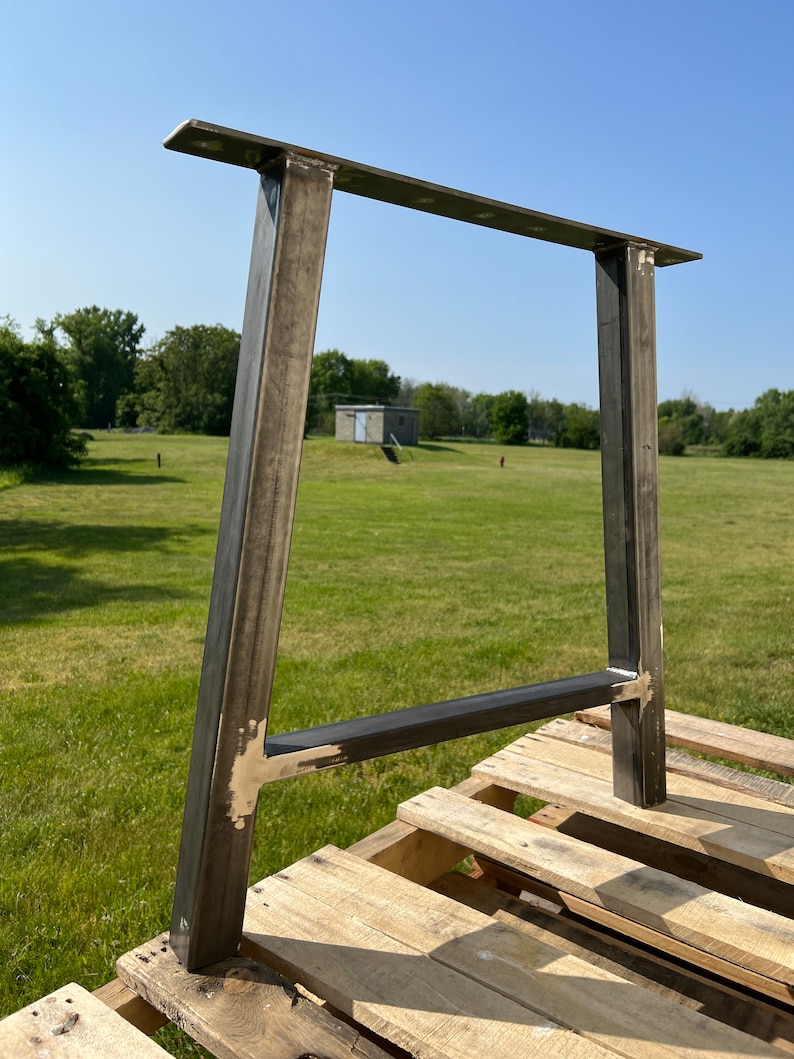Puede incluir: Una pata de mesa de metal con una parte superior rectangular y dos soportes verticales. La pata est&aacute; hecha de acero y tiene un acabado gris oscuro. La pata est&aacute; sentada sobre una plataforma de madera.