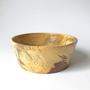 May include: A small, wooden bowl with a light brown and dark brown wood grain pattern. The bowl is slightly flared at the top and has a rounded bottom.