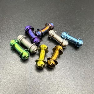 May include: A collection of colorful metal objects, each with a cylindrical body and flared ends. The objects are in shades of green, silver, purple, gold, and blue. The items are scattered on a dark surface.