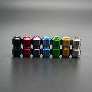 May include: A collection of seven small, cylindrical objects in various metallic colors: black, red, purple, blue, green, gold, and silver. Each object has a two-part design with a slightly wider base and top.