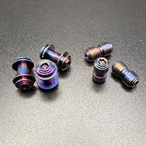 May include: An assortment of iridescent metal hardware components, including bolts and cylindrical pieces. The bolts have a tiered design, while the cylinders feature a hex socket. The color palette shifts between blues, purples, and golds.