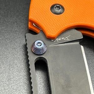 May include: Close-up of a folding knife with an orange handle and a black blade. The blade has a small inscription and a textured, blue-toned pivot screw. The knife is partially open, revealing the sharp edge.