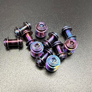 May include: A collection of iridescent, cylindrical plugs with a flared design. The plugs display a range of colors, including purple, blue, and gold. The jewelry is made of metal and has a screw-on design.