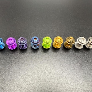 May include: A row of eight colorful metal plugs in various shades, including blue, purple, green, and gold. The plugs have a cylindrical shape with a flared base and a textured surface. They are arranged in a line against a dark background.