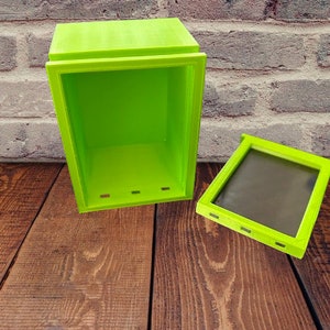 May include: A bright green box with a hinged lid, open to reveal an empty interior. The box is made of plastic and has a smooth surface. The box is sitting on a wooden surface.