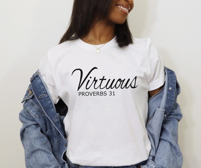 May include: White t-shirt with black text that reads "Virtuous Proverbs 31". The text is in a cursive font. A person is wearing the t-shirt and a denim jacket.