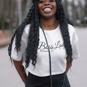 May include: A woman wearing a white cropped t-shirt with the text "Boss Lady Proverbs 31" in black script. She is also wearing black pants and a black hat.