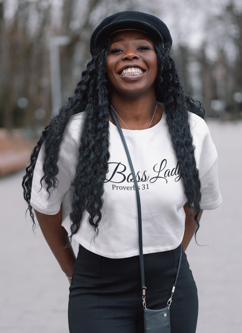 May include: A woman wearing a white cropped t-shirt with the text "Boss Lady Proverbs 31" in black script. She is also wearing black pants and a black hat.