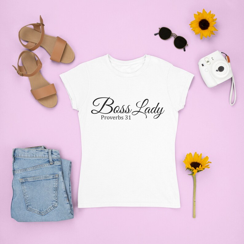 May include: White t-shirt with the text "Boss Lady Proverbs 31" in black script font. The shirt is laid flat on a pink surface with a pair of brown sandals, a pair of sunglasses, a small camera, and two yellow sunflowers.