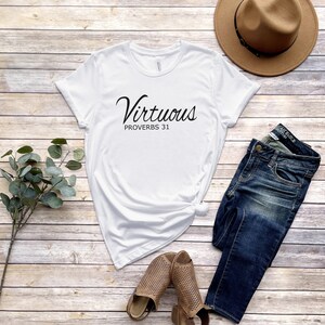 May include: White t-shirt with the text "Virtuous Proverbs 31" in black script font. The shirt is laid out on a woodgrain surface with a pair of brown sandals, blue jeans, and a brown fedora hat.