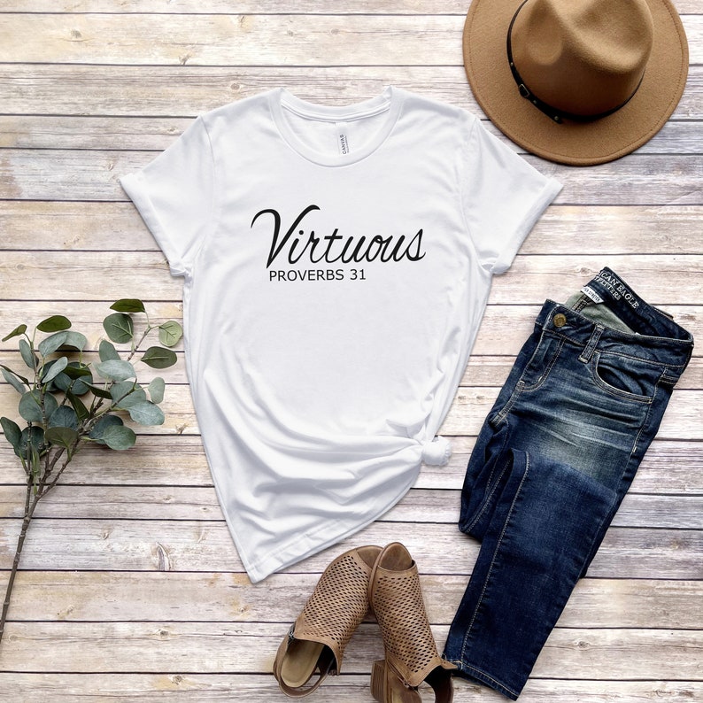 May include: White t-shirt with the text "Virtuous Proverbs 31" in black script font. The shirt is laid out on a woodgrain surface with a pair of brown sandals, blue jeans, and a brown fedora hat.