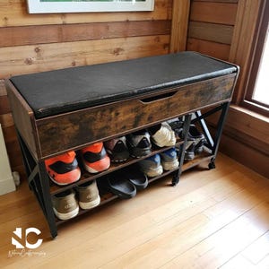 May include: A wooden shoe bench with a black cushioned top and two shelves for shoe storage. The bench has a dark brown finish with black metal legs and is filled with various shoes. The bench is 48 inches long.