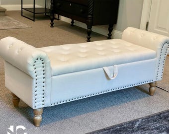 May include: A cream-colored storage bench with a tufted seat and rolled arms. The bench features decorative nailhead trim along the edges and sits on light-colored wooden legs. A handle is visible on the front.