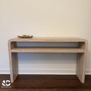 May include: A beige travertine console table with a smooth, rounded waterfall edge design and a horizontal middle shelf. The table features a natural stone texture with light brown veining throughout its surface and a small gold tray on top.
