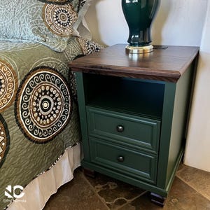 May include: A dark green nightstand with two drawers and an open storage shelf. The nightstand features a dark brown wooden top surface, small dark brown feet, and simple round black knobs on each drawer front. A dark green lamp sits on top.