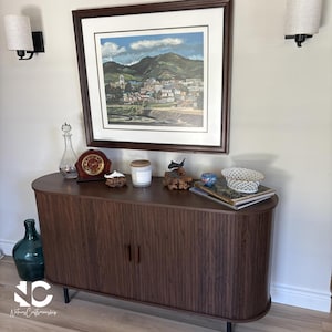 May include: A dark brown wooden cabinet with a curved front and vertical ribbed detailing. The cabinet has two doors and black legs. A framed landscape painting hangs above, with various decorative items on top, including a clock and candle.