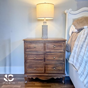 May include: A wooden nightstand with six drawers and brass knobs. A beige lamp with a cylindrical base and a lampshade sits on top. The nightstand is next to a bed with a white headboard and a patterned blanket.