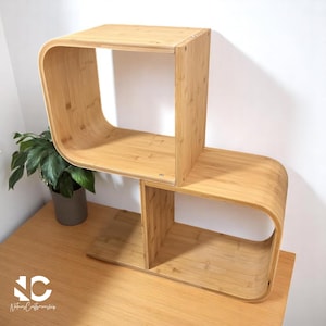 May include: A two-tiered bamboo shelf unit with a unique curved design. The light brown shelves are stacked, with one section turned 90 degrees. The unit is on a wooden surface, with a potted plant visible in the background.