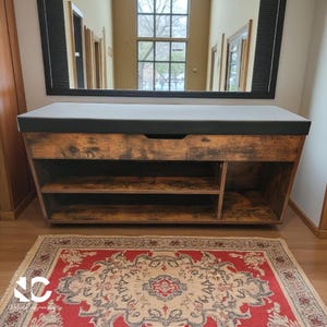 May include: A wooden entryway bench with a dark gray cushioned top and open shelving. The bench has a distressed wood finish and a built-in drawer. A decorative rug with a red and cream pattern is in front of the bench.