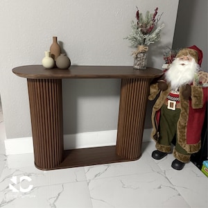 May include: A brown wooden console table with a rounded top and ribbed legs. Three decorative vases in neutral tones sit on the table. A Santa Claus figure stands to the right of the table. The table is approximately 48 inches long.
