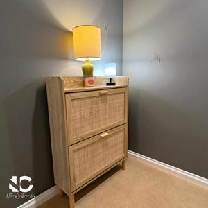 May include: A light wood-tone shoe cabinet with two woven rattan doors and wooden handles. A lamp with a yellow lampshade and a small green lamp sit on top. The cabinet is in a corner with gray walls and a beige carpet.