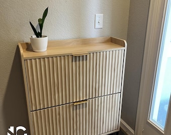 May include: A light-colored wooden cabinet with two drawers and a top shelf. The cabinet has a ribbed texture on the drawer fronts and gold-colored handles. A small white pot with a green plant sits on top. The cabinet is on a black metal frame.