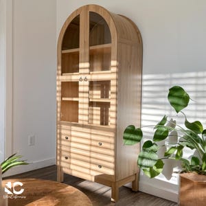 May include: A tall, light-colored wooden cabinet with an arched top and glass doors. The cabinet has shelves, drawers, and a natural wood finish. The cabinet is standing on a wooden floor.