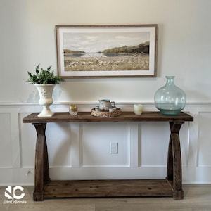 May include: A dark brown wooden console table with an X-frame base and a lower shelf. On top sits a white urn with greenery, a small glass, a woven tray with a pitcher and cups, a candle, and a large light blue glass vase. A landscape print hangs above.