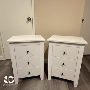 May include: Two white nightstands with three drawers each. The drawers have black knobs. The nightstands are rectangular with a simple design. The nightstands are placed on a wooden floor.