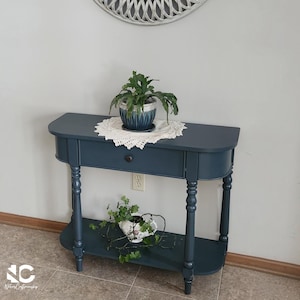 May include: A dark blue semi-circular console table with a single drawer, a lower shelf, and turned legs. A white crocheted doily sits on the top surface under a green plant in a blue pot, while a second green plant sits in a white pot below.