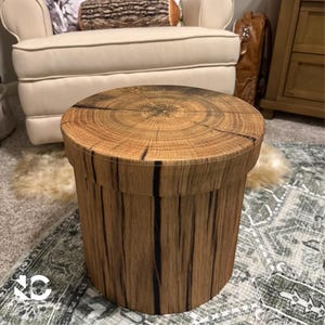 May include: A cylindrical wooden side table with a natural wood grain pattern. The table has a round top and a vertical grain design on the sides. The wood has a light brown color with dark brown accents. The table is approximately 18 inches tall.