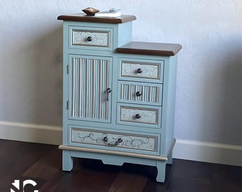 May include: A light blue wooden cabinet with a dark brown top and decorative drawer pulls. The cabinet has a combination of drawers and a door with vertical silver accents. The cabinet is on a dark wood floor.