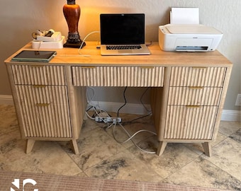 May include: A light wood desk with a ribbed drawer design and gold-toned hardware. The desk has a laptop, printer, and lamp on top. The desk has three drawers on the right and two drawers on the left. The desk is on a tiled floor.