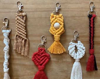 May include: Six colorful macrame keychains with different designs and colors, including red, white, yellow, and brown. Each keychain has a silver metal ring and a tassel.
