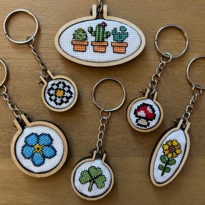 May include: A selection of handmade cross-stitch keyrings with diverse designs. The keyrings showcase a cactus garden, a mushroom, a sunflower, a four-leaf clover, a forget-me-not flower, and geometric patterns. Each design is set in a small wooden hoop.