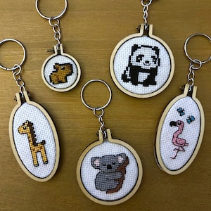 May include: Five keychains featuring miniature cross-stitch embroidery designs set in small wooden hoops. The designs include a yellow giraffe, a brown animal, a black and white panda, a grey koala, and a pink flamingo with two blue butterflies.