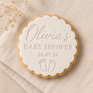 Peut inclure: Un biscuit rond festonn&eacute; recouvert d'un gla&ccedil;age blanc avec le texte en relief Olivia's BABY SHOWER 26.07.26 et une paire d'empreintes de pieds de b&eacute;b&eacute;. Le biscuit pr&eacute;sente un bord dor&eacute; clair et une surface lisse avec des lettres nettes.